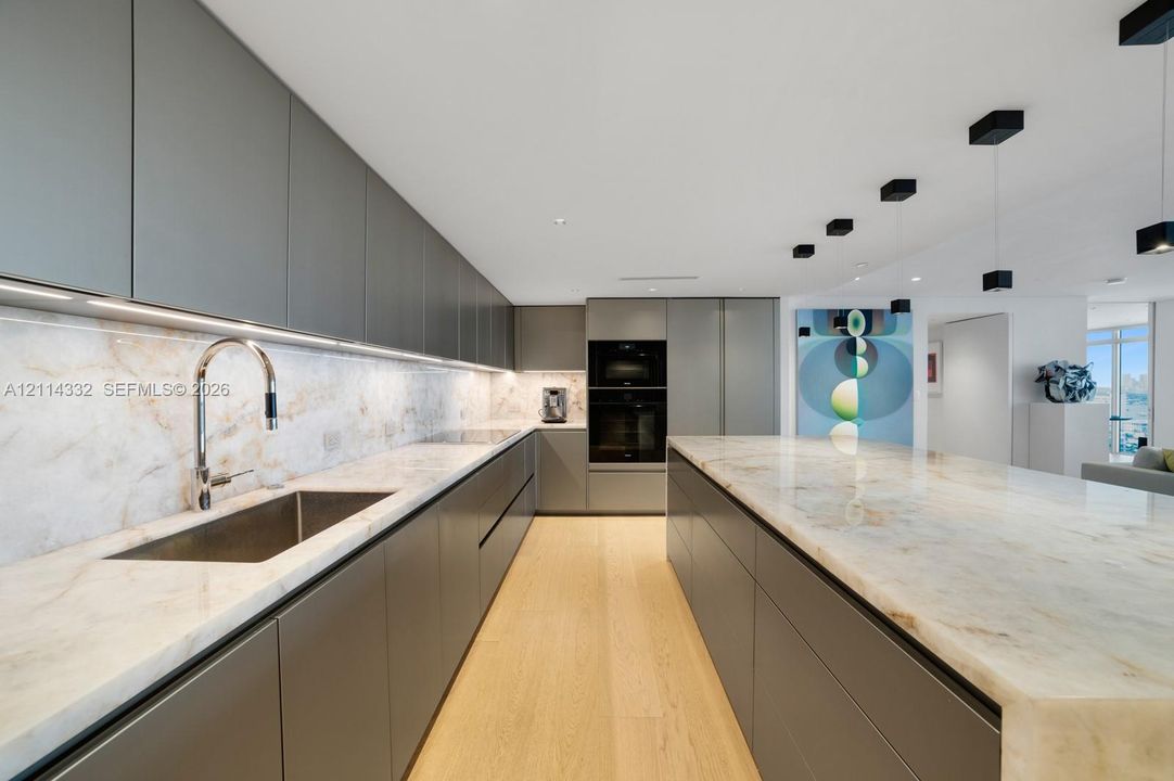 Sleek, handleless grey cabinetry, stone countertops with waterfall edge island, under-cabinet lighting, stainless steel undermount sink with arched faucet, and wood-finish flooring