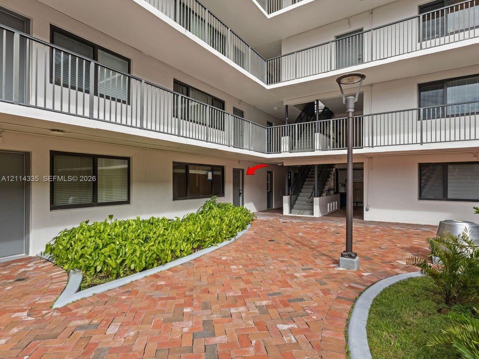 Courtyard featuring herringbone-patterned brick pavers, integrated landscape beds, tiered structural metal balconies, and a built-in metal light fixture