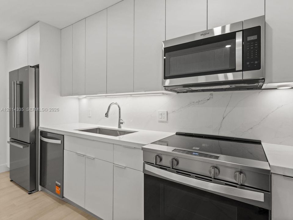 Kitchen featuring flat-panel white cabinetry, stone-patterned backsplash, stainless steel appliances, and integrated under-cabinet lighting