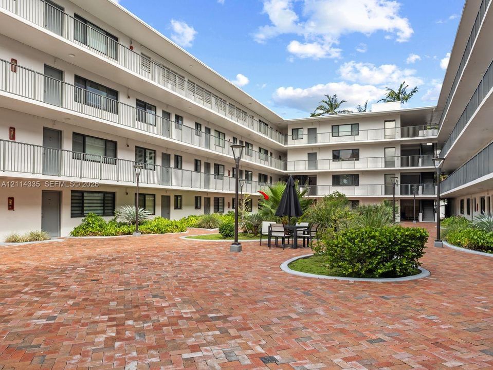 Courtyard featuring herringbone-pattern brick paving, multi-story building exterior with metal railings, and integrated landscaping with manicured shrubs