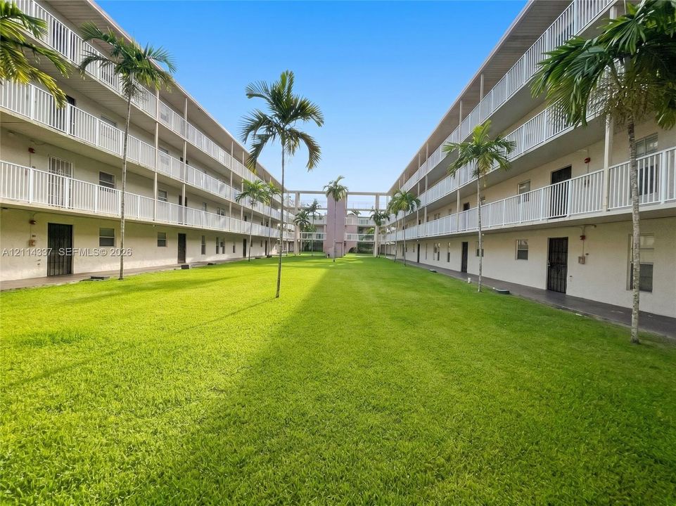 Multi-story residential complex featuring white metal balcony railings, a central grassy courtyard, and mature palm tree landscaping