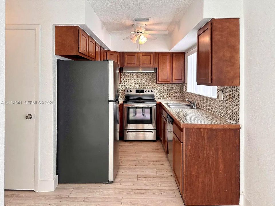 Kitchen featuring wood cabinetry, stainless steel appliances, mosaic tile backsplash, wood-finish flooring, and a ceiling-mounted light fixture