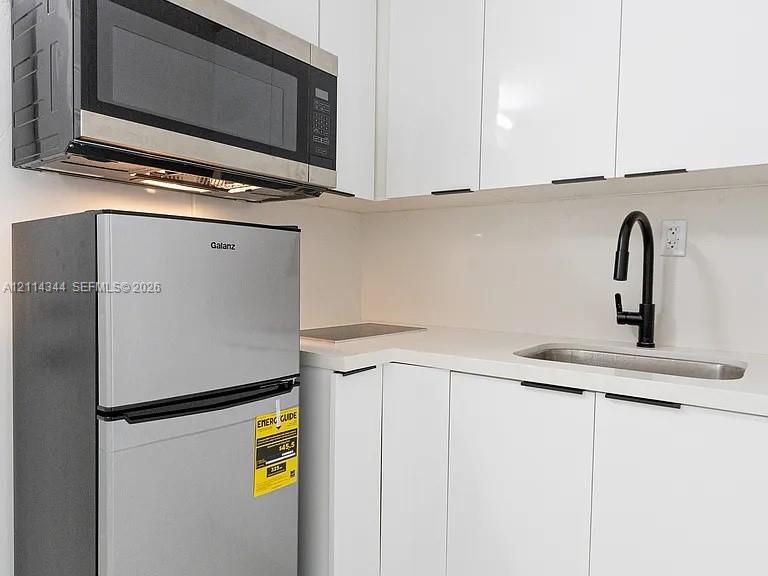 High-gloss white cabinetry featuring matte black hardware, stainless steel refrigerator, stainless steel over-the-range microwave, solid-surface countertop with integrated sink, and matte black high-arc faucet