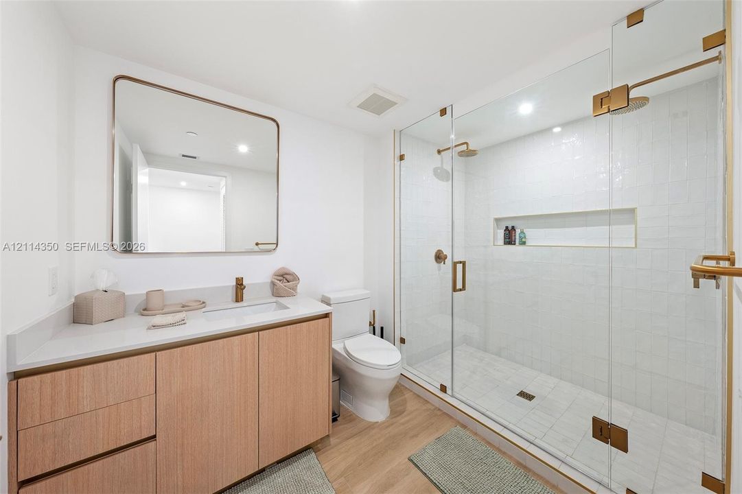 Bathroom featuring a vanity with light wood cabinetry and a solid-surface countertop, a rectangular mirror with a metallic frame, a walk-in shower with a glass enclosure and built-in recessed shelving, and metallic hardware throughout