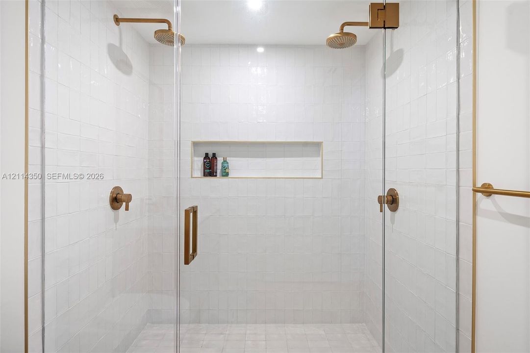 Walk-in shower featuring white wall tiling, dual brass-finish rainfall showerheads, a recessed storage niche, and a frameless glass enclosure with brass hardware