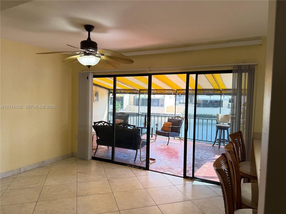 Interior featuring light-toned tile flooring, ceiling fan with integrated lighting, and large sliding glass doors opening to a covered patio with paved surfacing and an awning