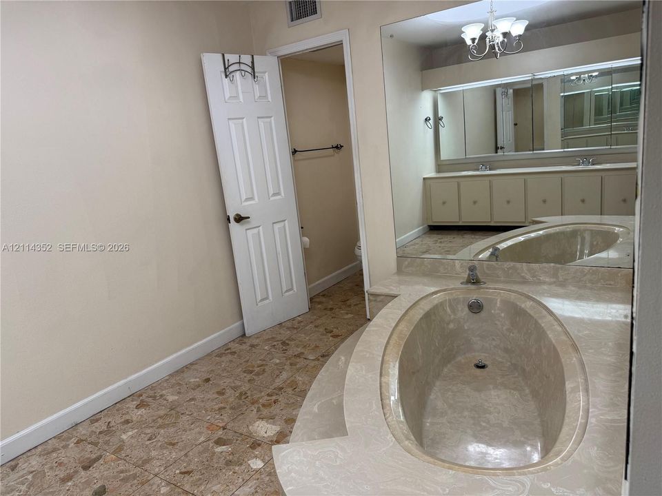 Bathroom featuring a dual-sink vanity with cabinetry, expansive wall mirror, drop-in soaking tub with stone-finish surround, and stone-tile flooring