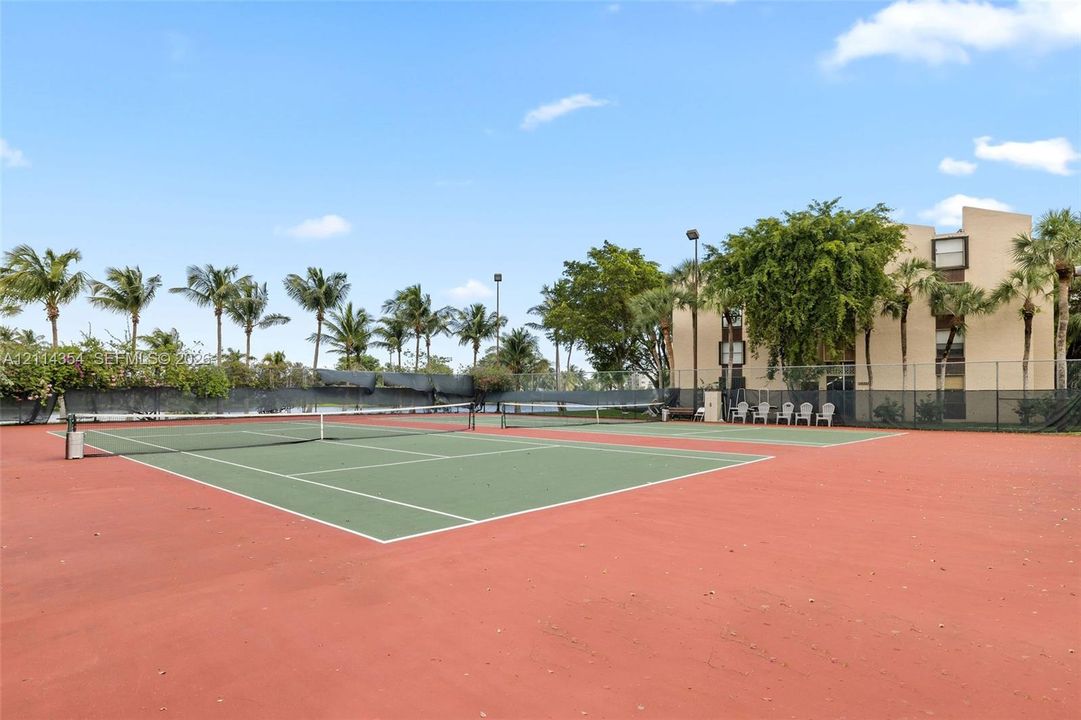 Hard-court tennis facility featuring perimeter chain-link fencing, dual courts, overhead lighting, and mature palm landscaping