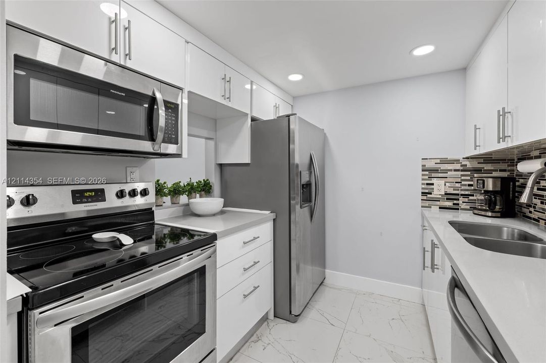 Kitchen featuring white gloss cabinetry, stone-finish countertops, stainless steel appliances, a multi-tone glass tile backsplash, and large-format porcelain tile flooring
