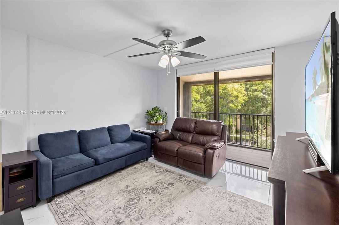 Living area featuring polished tile flooring, a multi-blade ceiling fan with integrated lighting, and full-width sliding glass doors opening to a balcony with metal railing
