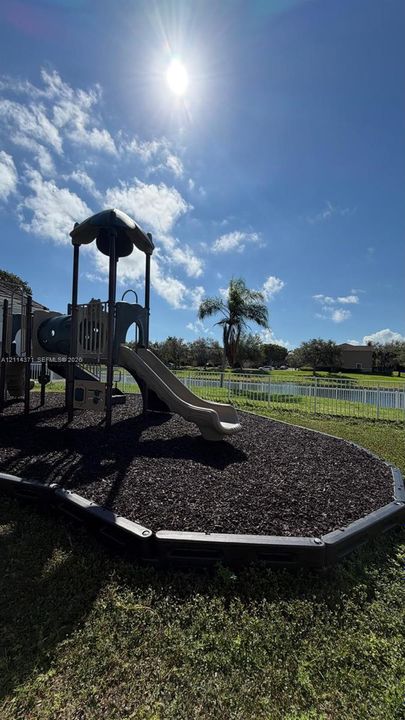 Playground structure featuring a slide, climbing platform, mulch surfacing, and a defined border, situated in a grass-covered backyard with a perimeter fence
