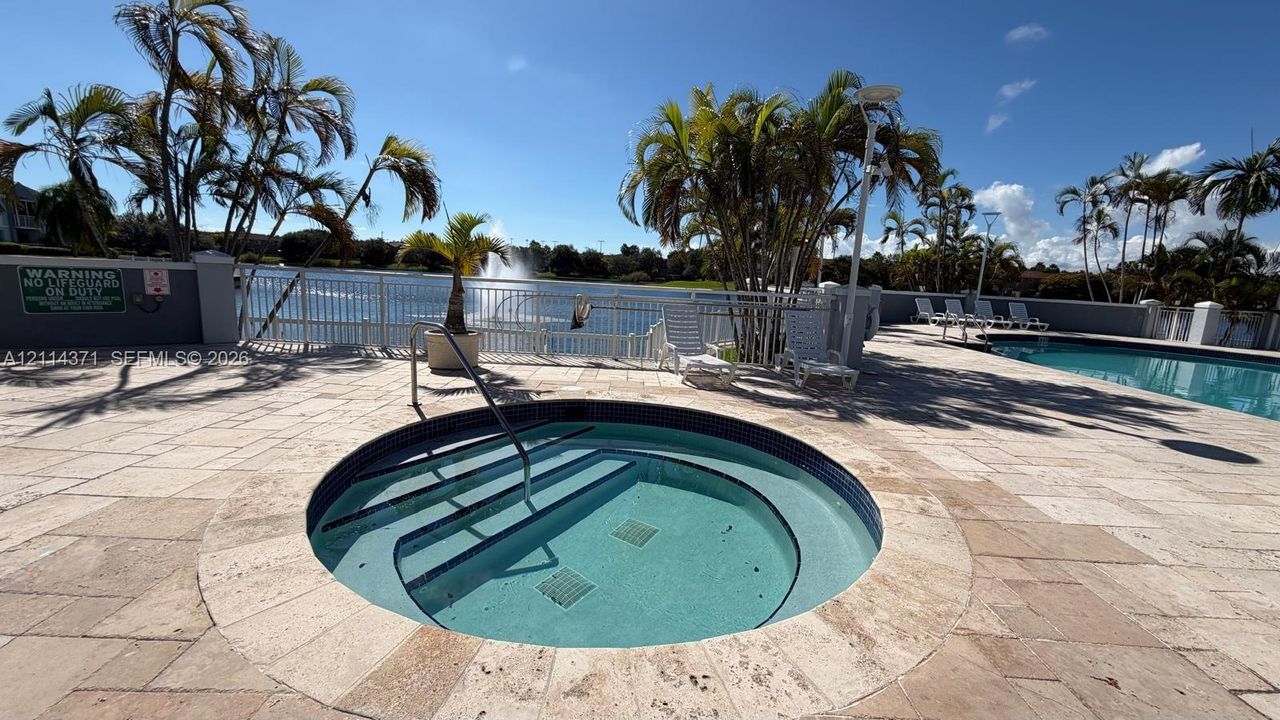 Circular in-ground spa featuring stone coping, surrounded by a paved deck, swimming pool, and water feature backdrop