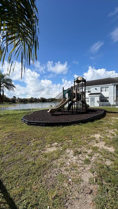 Outdoor recreational area featuring a playground structure with slides, a mulched surface with a composite border, and a metal perimeter fence overlooking a pond