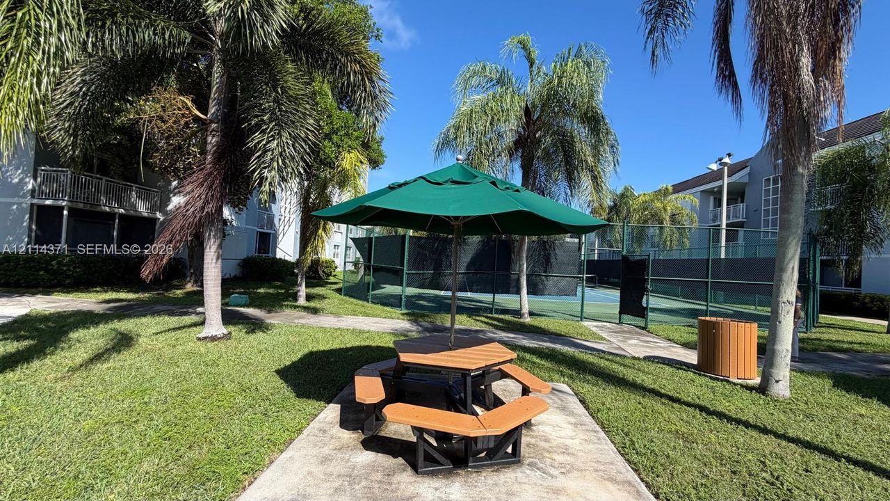 Outdoor common area featuring a hardscaped patio, manicured lawn, mature palms, and an enclosed tennis court