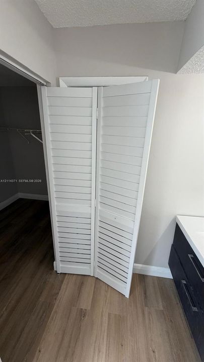 White louvered bifold doors, wood-finish flooring, and a dark-toned vanity featuring a light-colored countertop