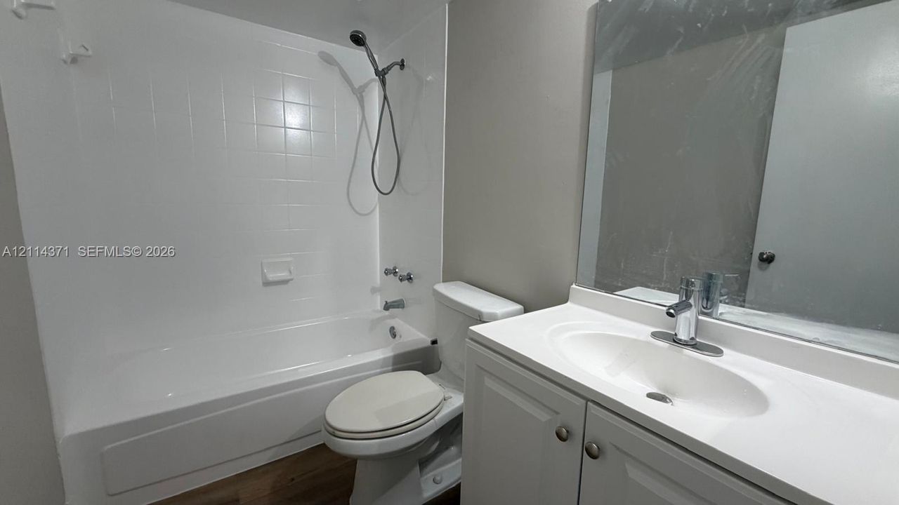 Bathroom featuring a white vanity cabinet with a integrated sink, large wall-mounted mirror, white acrylic tub-shower combination with a handheld showerhead, and wood-finish flooring
