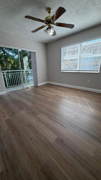 Wood-finish flooring, sliding glass door with balcony access, multi-pane window with horizontal blinds, and a five-blade ceiling fan with integrated light fixture