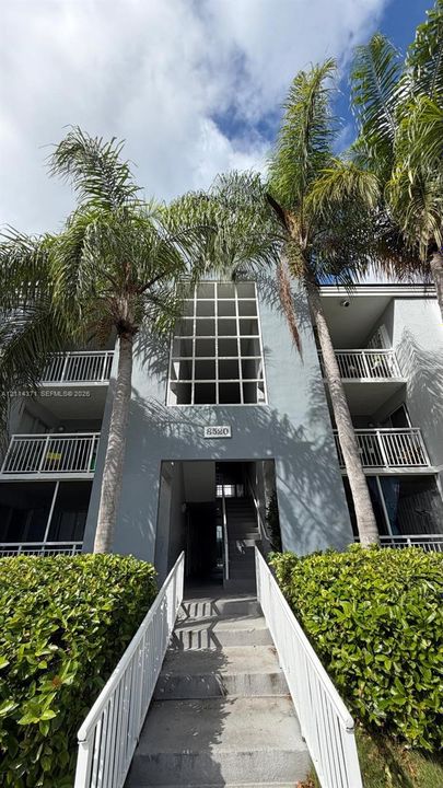 Multi-story stucco exterior featuring stacked balconies with white metal railings, a central grid-patterned window, and a concrete walkway with white handrails