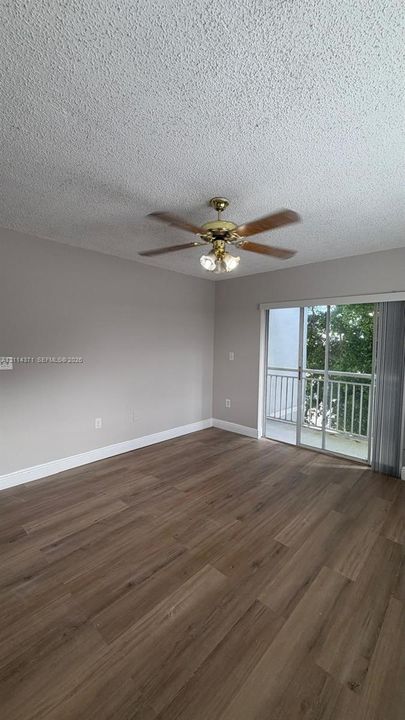 Interior room featuring wood-finish flooring, textured ceiling, ceiling fan with integrated lighting, and a sliding glass door providing access to a balcony with railings