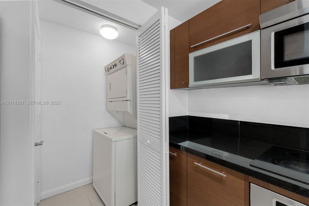 Laundry closet with louvered folding doors, featuring stacked washer and dryer, adjacent to a kitchen with wood-finish cabinetry, dark stone-style countertops, and stainless steel built-in microwave