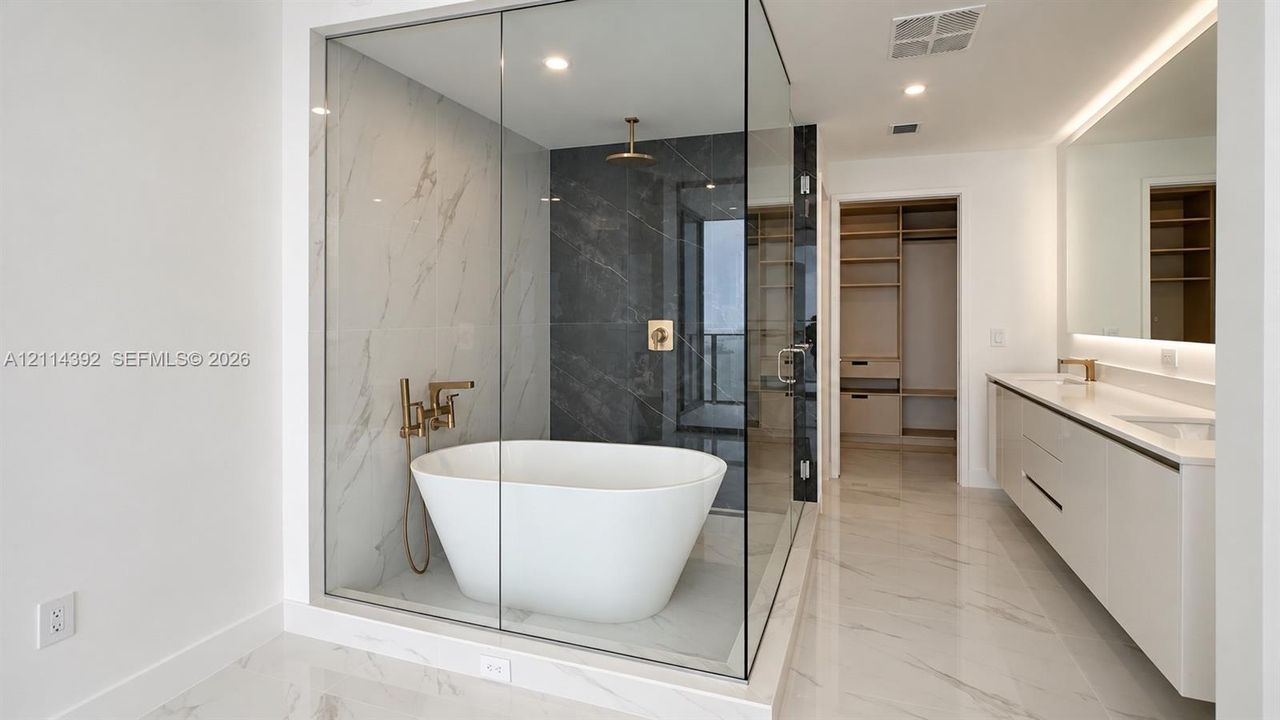 Glass-enclosed wet room featuring a freestanding soaking tub, dual-tone tile surround, and brushed gold fixtures, complemented by a dual-sink vanity with under-cabinet lighting and a walk-in dressing area with built-in shelving