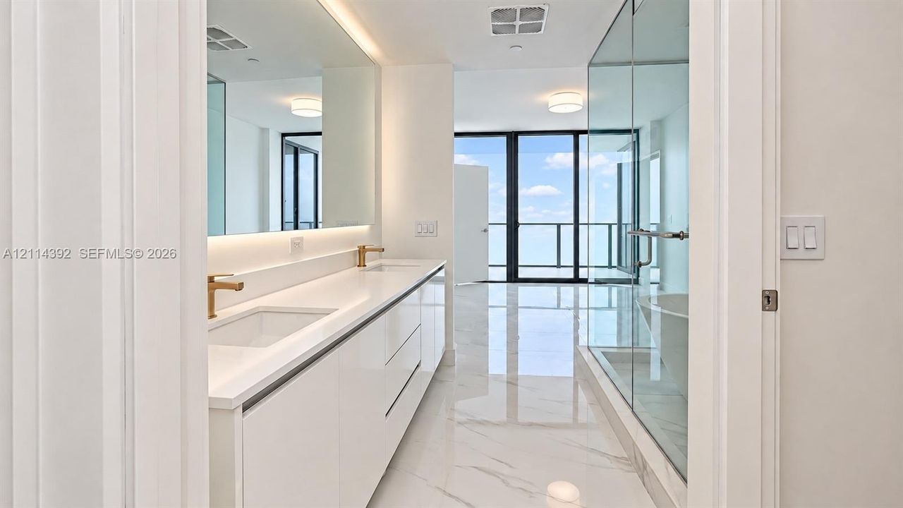 Bathroom featuring white cabinetry, dual undermount sinks with brass-tone fixtures, wall-to-wall mirror, large-format tile flooring, and a glass-enclosed shower