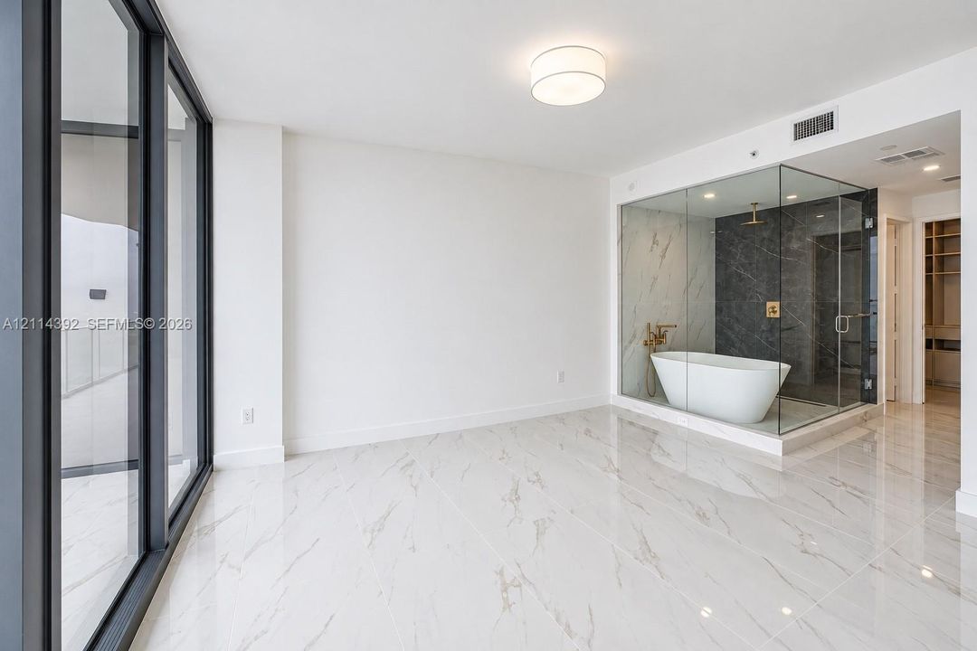 Floor-to-ceiling glass paneling, polished stone-finish flooring, glass-enclosed shower, freestanding soaking tub, and gold-tone fixtures