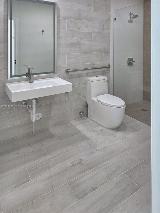 Wall-mounted rectangular basin with a single-handle faucet, full-height tile wall surfacing, walk-in glass shower enclosure, elongated one-piece toilet, and wood-finish tile flooring