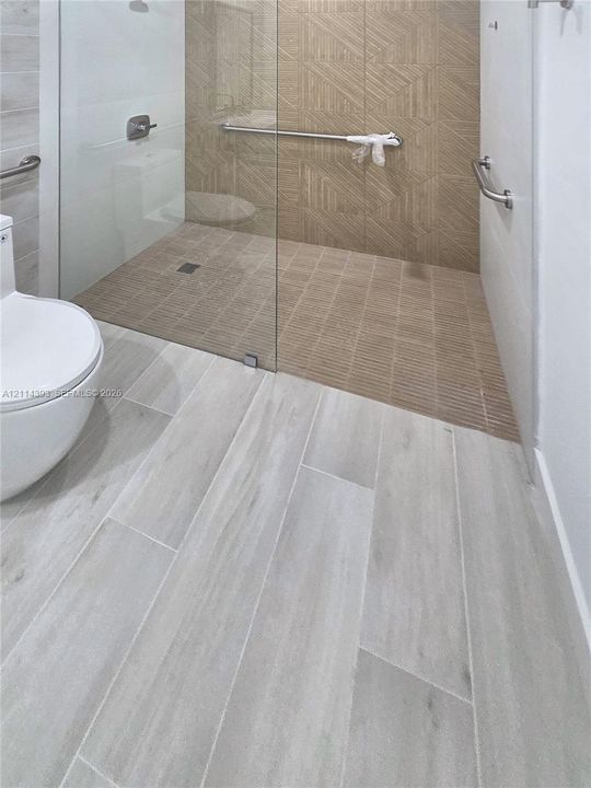 Walk-in shower featuring a frameless glass enclosure, geometric-patterned tile wall accents, wood-finish floor tiling, and wall-mounted metal support bars
