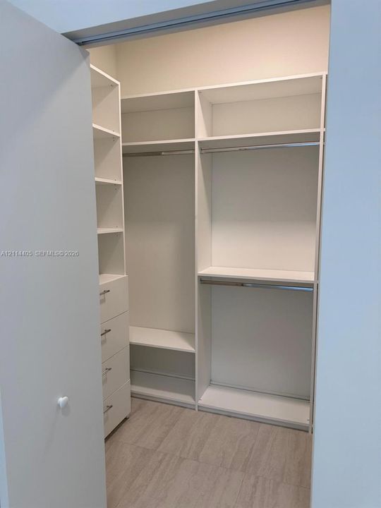 Custom storage system featuring white built-in cabinetry, multiple adjustable shelves, integrated hanging rods, and a four-drawer stack with metallic hardware, finished with wood-finish flooring