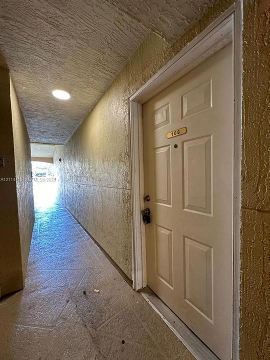 Six-panel entry door with brass-tone hardware and integrated peephole, featuring a textured stucco exterior wall and recessed ceiling lighting