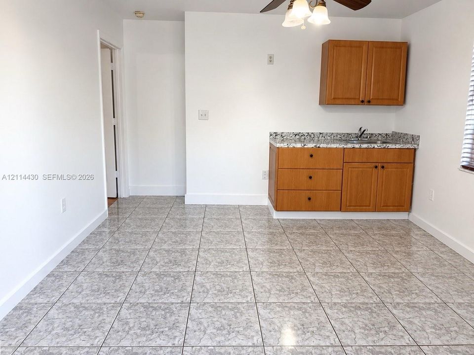 Kitchenette featuring wood-finish cabinetry, patterned stone-style countertops, integrated sink, neutral floor tiling, and a ceiling-mounted light fixture