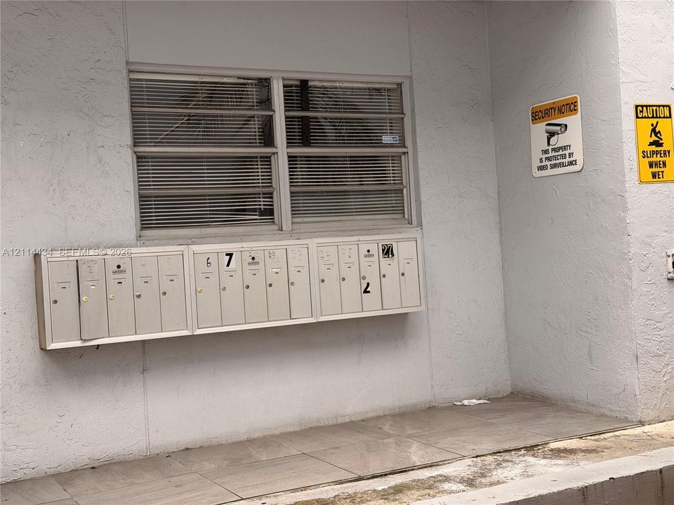 Textured exterior wall featuring a multi-unit wall-mounted metal mailbox bank, horizontal louvered window, and ceramic tile flooring