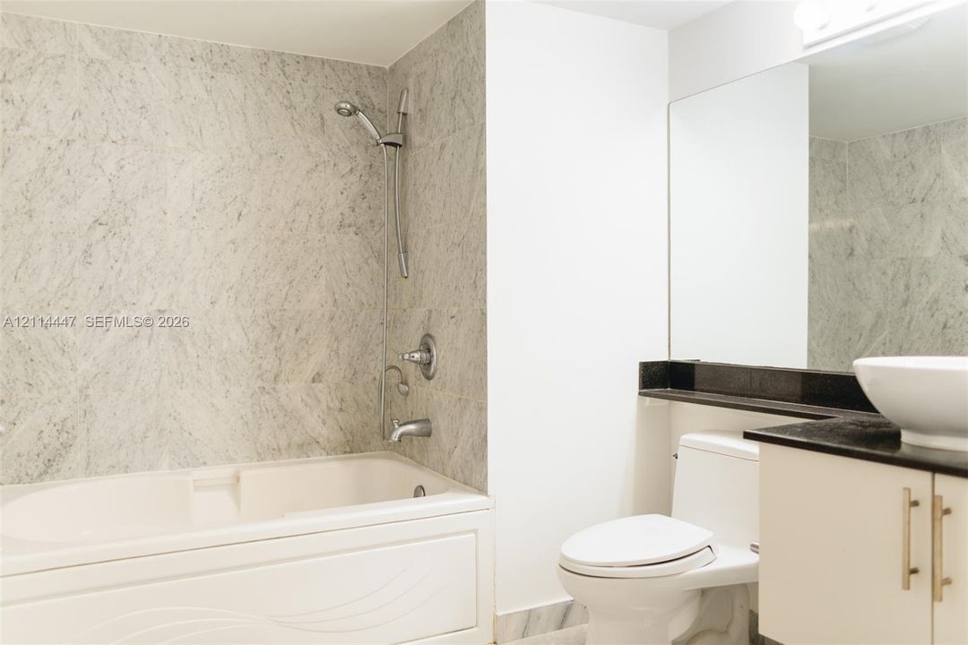 Bathroom featuring a deep-soaking tub with stone-patterned wall tile, an adjustable shower head, a vessel sink with a dark stone countertop, and a wide wall-mounted mirror