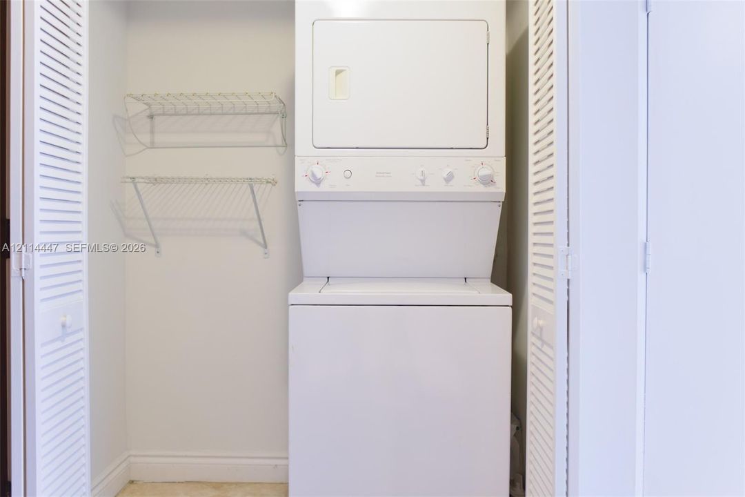 Laundry closet featuring stacked white appliance unit, bi-fold louvered doors, and dual-level wire shelving
