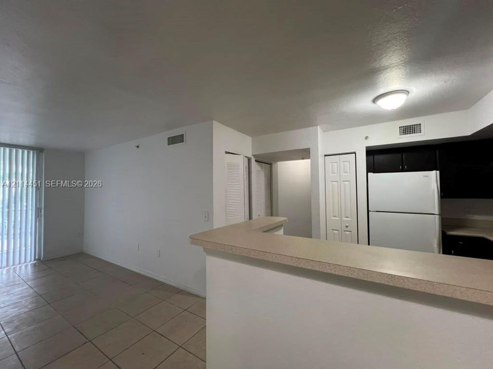 Living area featuring neutral tile flooring, kitchen with dark cabinetry, solid-surface breakfast bar, and vertical blind sliders