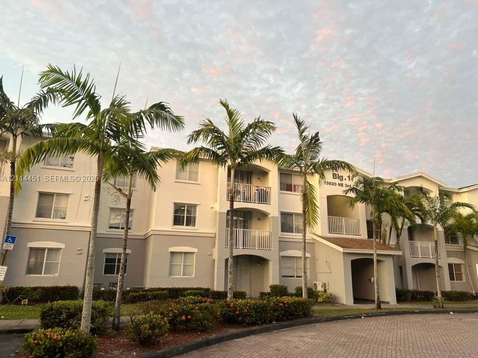 Multi-story residential building featuring a two-tone stucco exterior, covered private balconies with metal railings, and mature palm tree landscaping