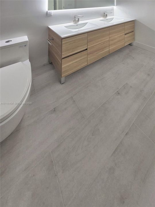 Modern floating vanity featuring light-toned cabinetry, dual rectangular under-mount sinks, integrated lighting, and neutral large-format tile flooring