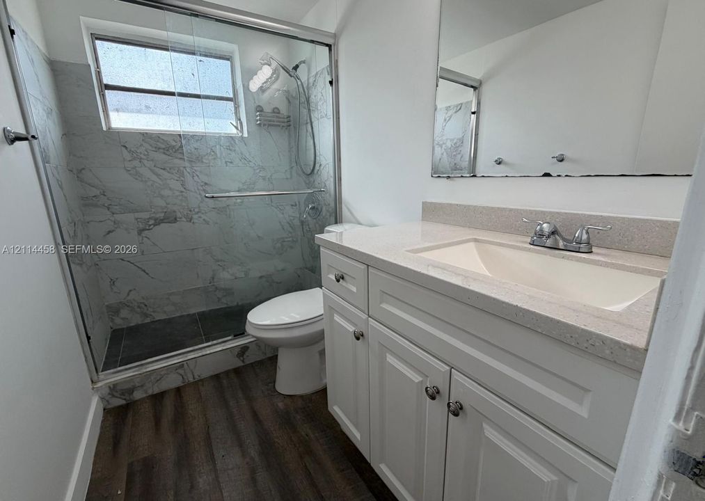 White vanity cabinet featuring a solid surface countertop with integrated basin, glass-enclosed shower with floor-to-ceiling tile, and wood-finish flooring