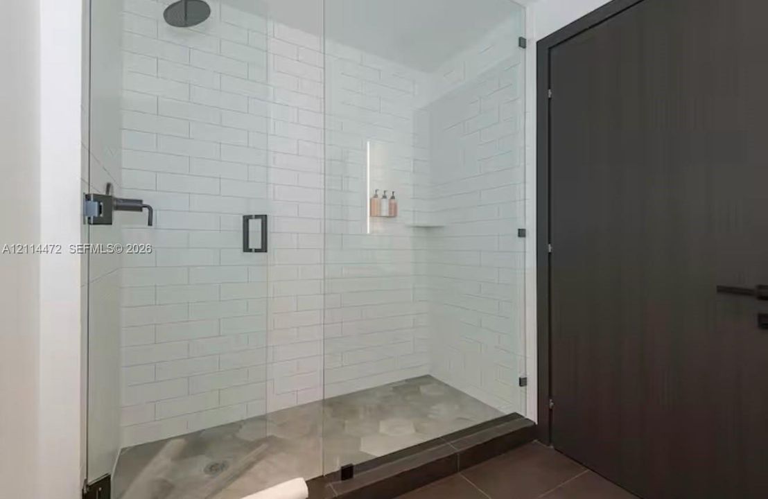 Walk-in shower featuring a frameless glass enclosure, white subway tile surround, hexagonal tile flooring, matte black rainfall showerhead, and integrated recessed shelving