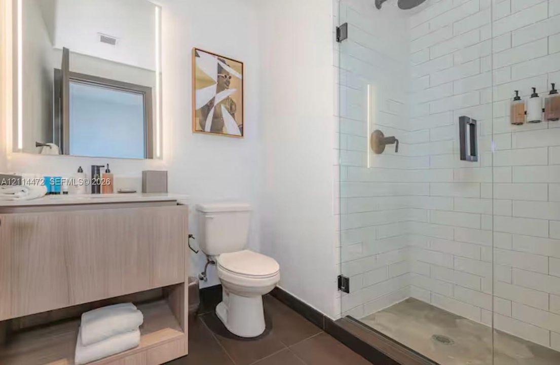 Wood-finish floating vanity with an integrated countertop, illuminated wall-mounted mirror, walk-in shower with white subway tile surround, and large-format tile flooring