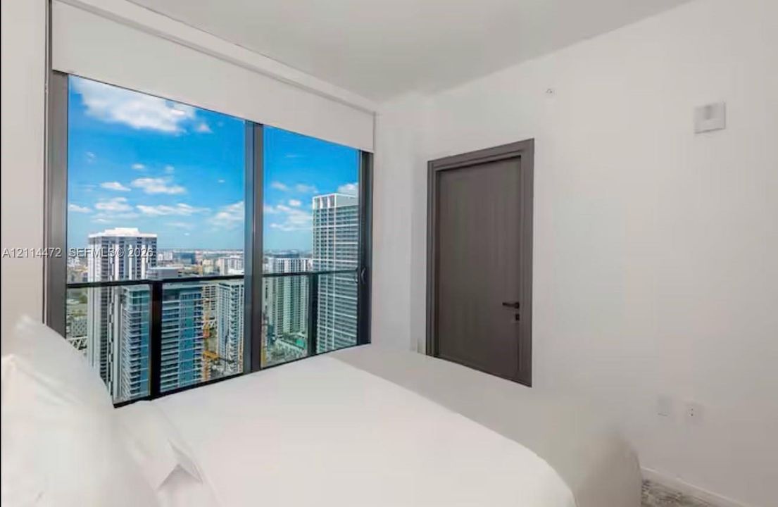 Floor-to-ceiling windows with dark metallic frames, panoramic cityscape, retractable roller shade, and solid-core door with matte black hardware