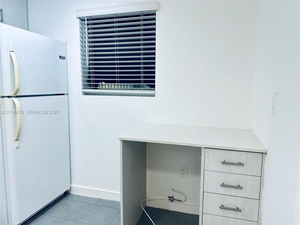 Kitchen area featuring a white top-mount refrigerator, light-toned built-in cabinetry with three drawers, a stone-composite countertop, and a window fitted with horizontal blinds