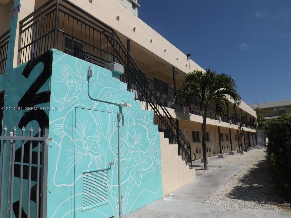Multi-story exterior featuring a custom wall mural, black metal railings, concrete walkway, and palm tree landscaping