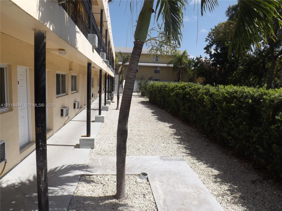 Exterior corridor featuring concrete walkways, dark metal support columns, gravel landscaping, and tall privacy hedging