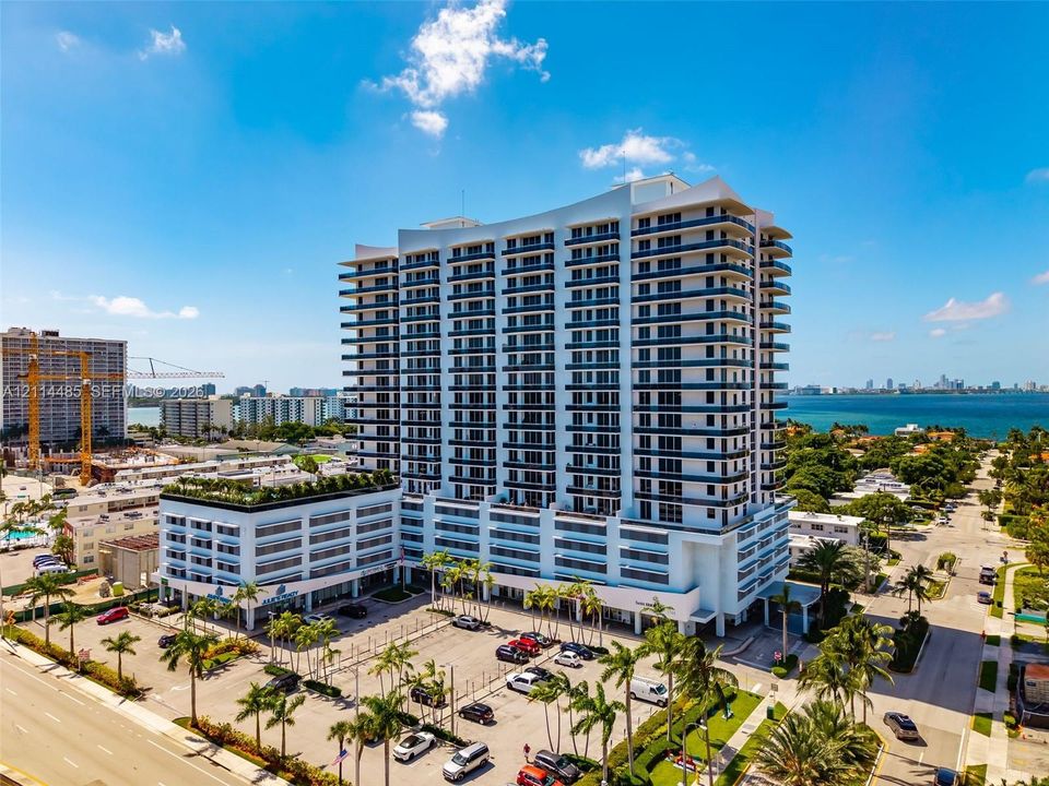 High-rise residential building featuring a curved architectural silhouette, wrap-around balconies, glass-paneled railings, and surface parking