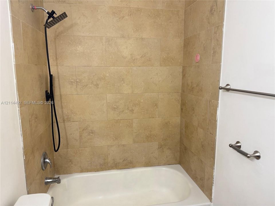 Bathtub surround featuring large-format neutral-tone tiling, a rectangular rainfall shower head, matte black hardware, and attached brushed-nickel fixtures