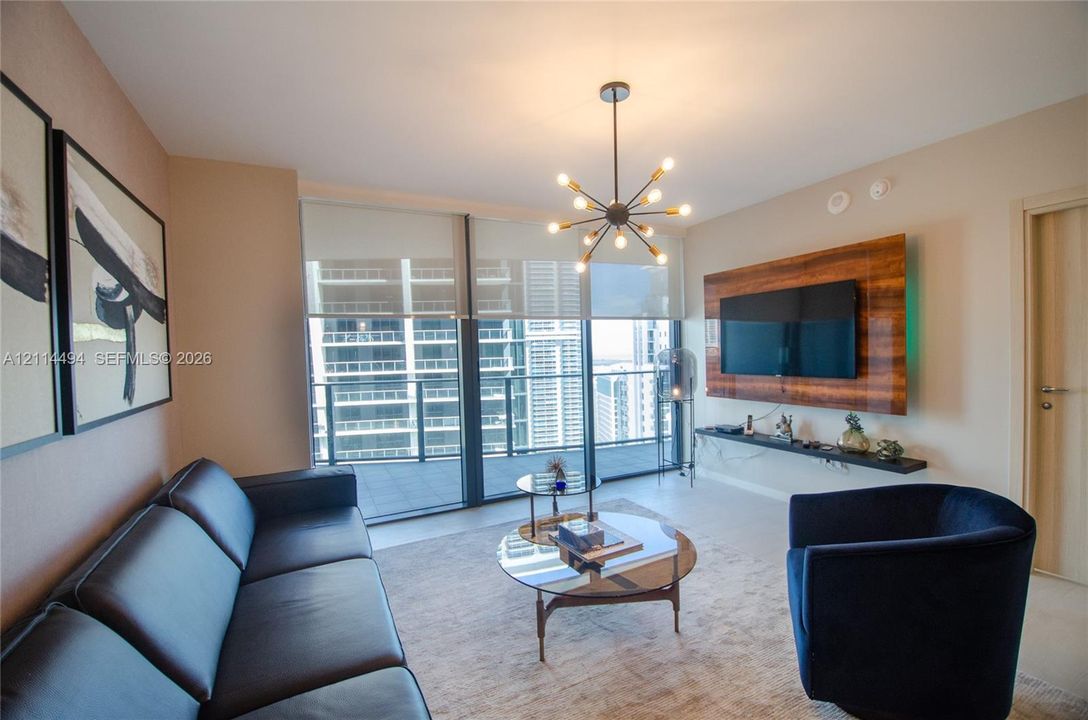 Floor-to-ceiling windows with integrated roller shades, wood-finished media wall with built-in shelving, multi-bulb modern sputnik chandelier, and tile flooring