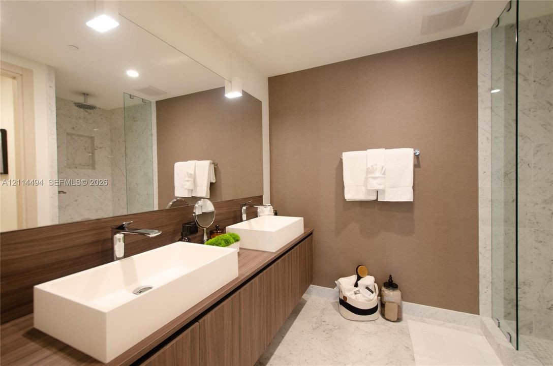 Floating vanity with dual rectangular vessel sinks, expansive wall-to-wall mirror, stone-finish flooring, and glass-enclosed shower with tiled surround