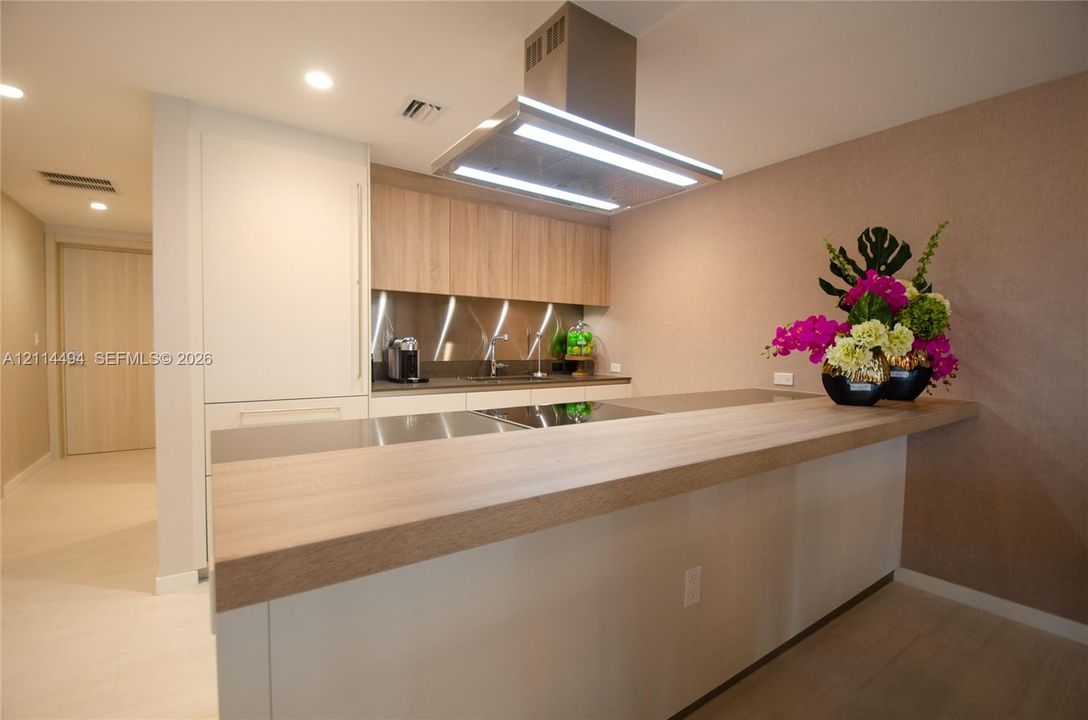 Modern kitchen featuring wood-finish cabinetry, sleek stainless steel backsplash, integrated cooktop, extended breakfast bar, and a stainless steel island-mounted range hood